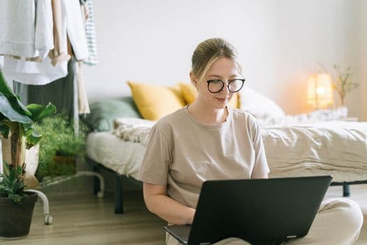 Caucasian woman wearing eyeglasses working on a laptop in a cozy bedroom setting.