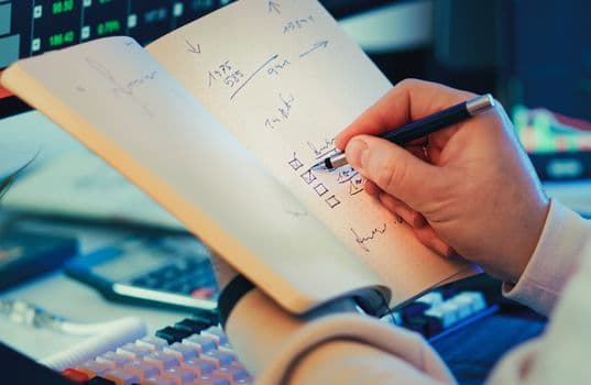 A hand writing in a notebook during stock market analysis on a desk.