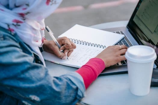 Crop anonymous Muslim woman student in casual clothes and headscarf sitting at table with cup of takeaway coffee and taking notes in notebook while browsing laptop in daytime