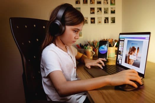 A focused young girl wearing headphones works on a creative project using a laptop and design software.