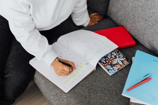 A woman in a white sweater writes notes on a notebook while studying on a sofa.