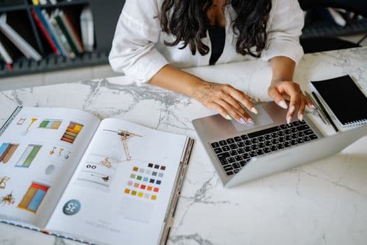 A woman works on a laptop at a marble desk with a design book and notebook nearby.