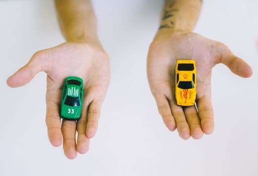 Pair of adult hands holding green and yellow toy cars on a white surface, symbolizing choice.
