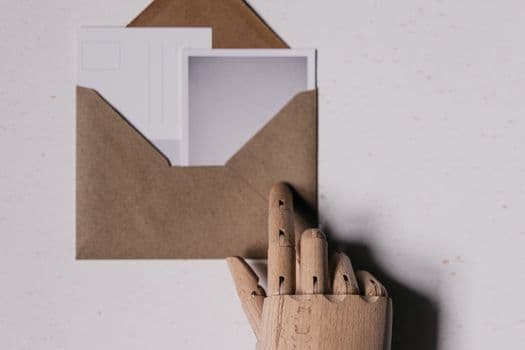 A wooden mannequin hand holds a brown envelope containing blank postcards.