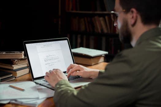 A man focuses on typing a document in a warm, cozy library environment, surrounded by books.