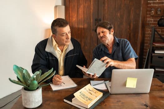 Two men working together on a creative project with books and a laptop.
