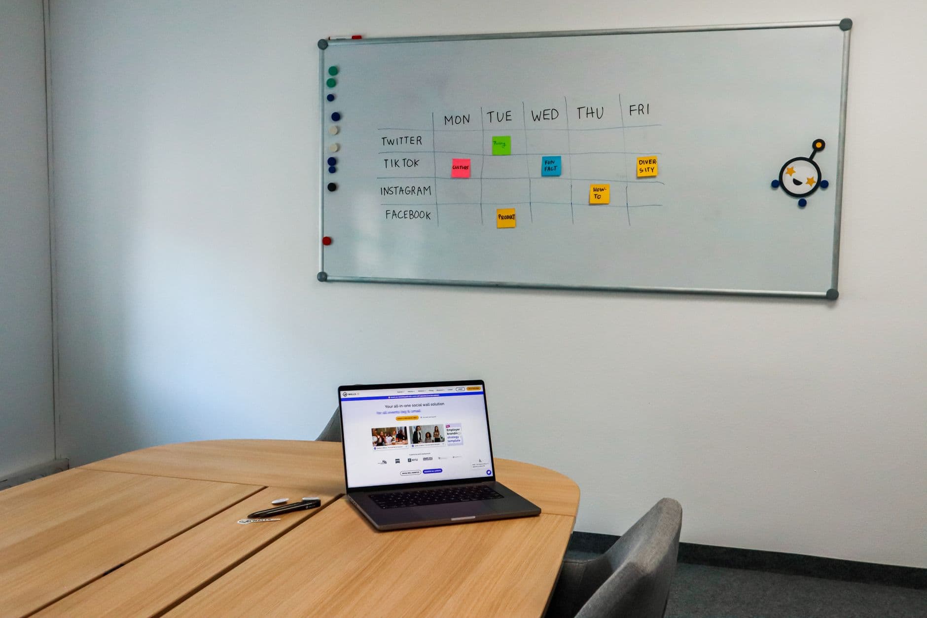Office setup featuring a conference table, laptop, and organized whiteboard schedule, highlighting a tech-driven workspace.