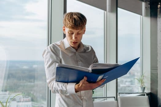 Businessman reading documents in an office with a city view, holding a blue binder.