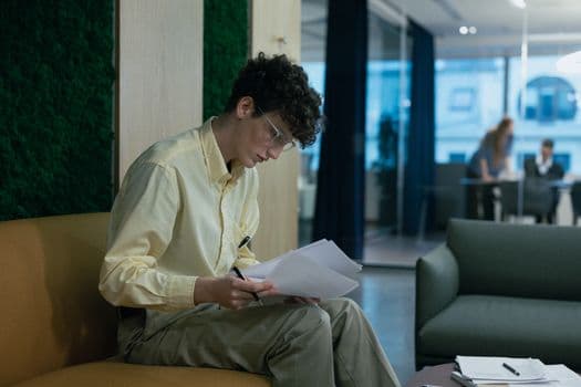 A focused young professional reading documents on a couch in a modern office environment.