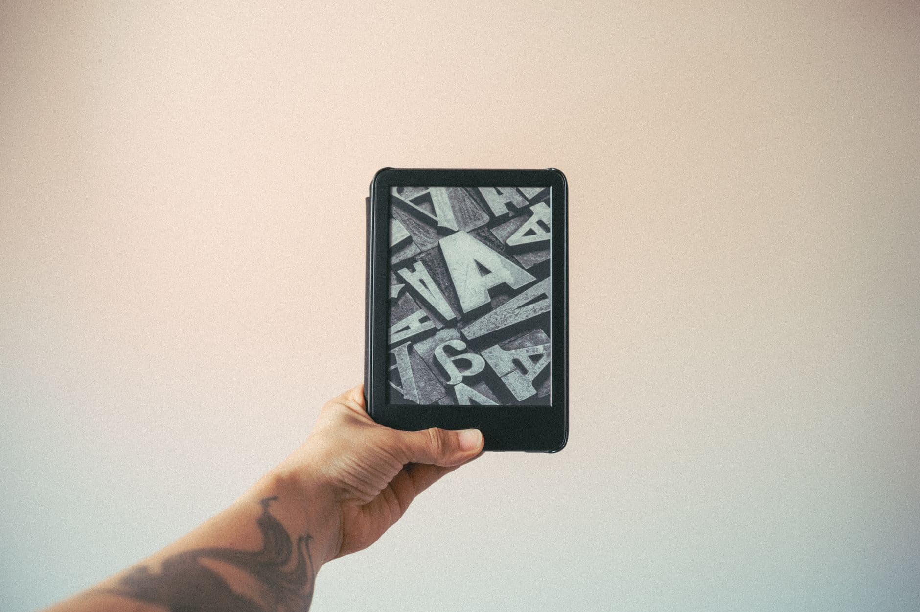 A tattooed hand holding an e-reader displaying typography design against a plain background.