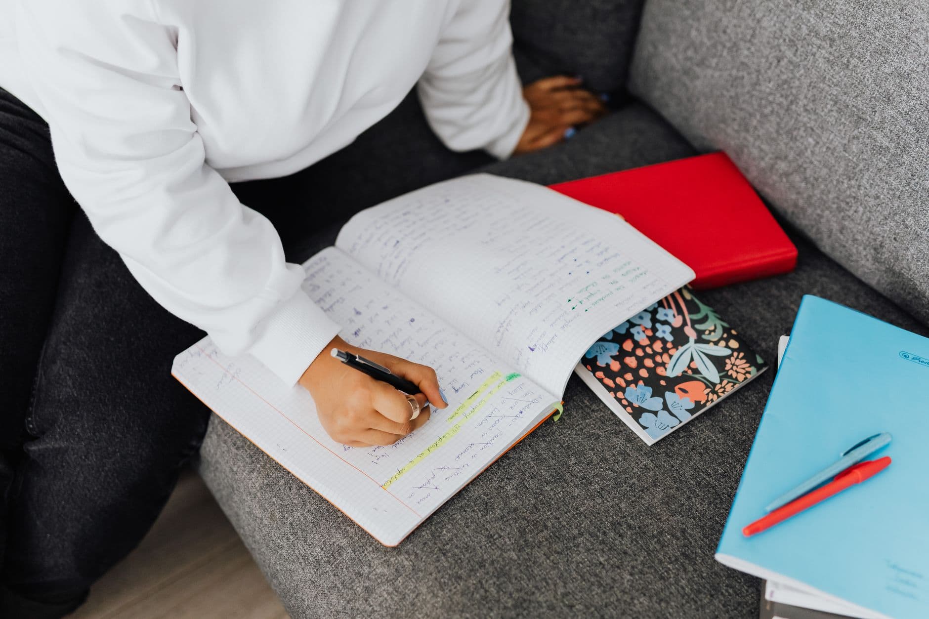 A woman in a white sweater writes notes on a notebook while studying on a sofa.