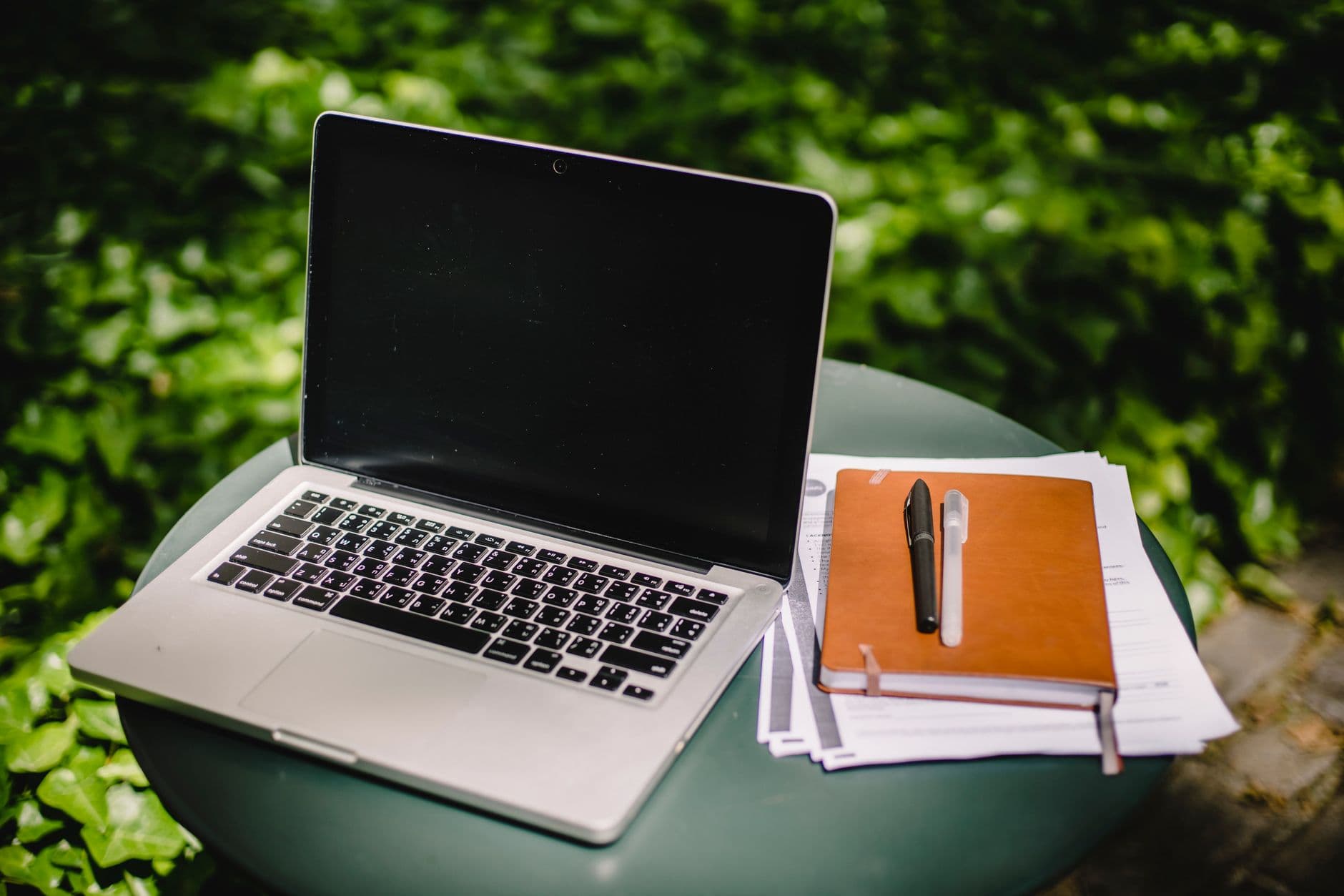 Modern netbook with empty screen placed near brown notebook and papers on round table against blurred green garden